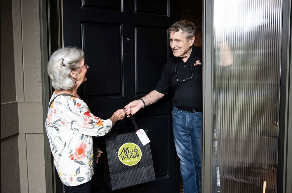 A woman hands a Meals on Wheels bag to a man.